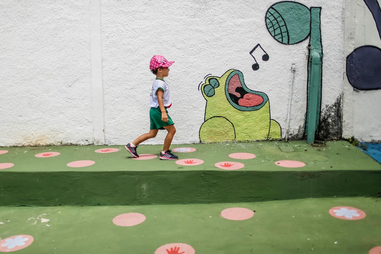Calçada pintada de verde e muro com pintura de sapo cantando. Criança caminha na frente do muro.