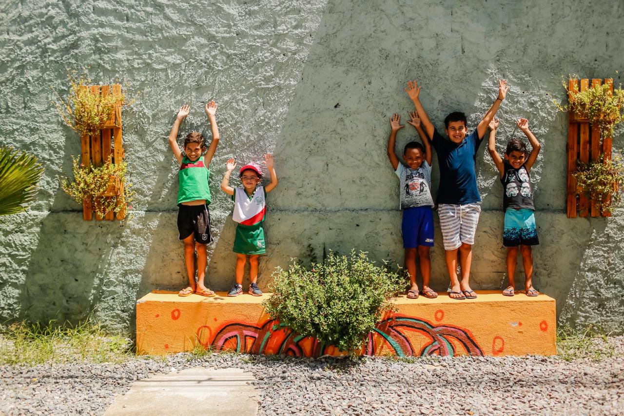 Cinco crianças sorridentes e de braços erguidos, encostadas em um muro revitalizado do bairro Vasco da Gama