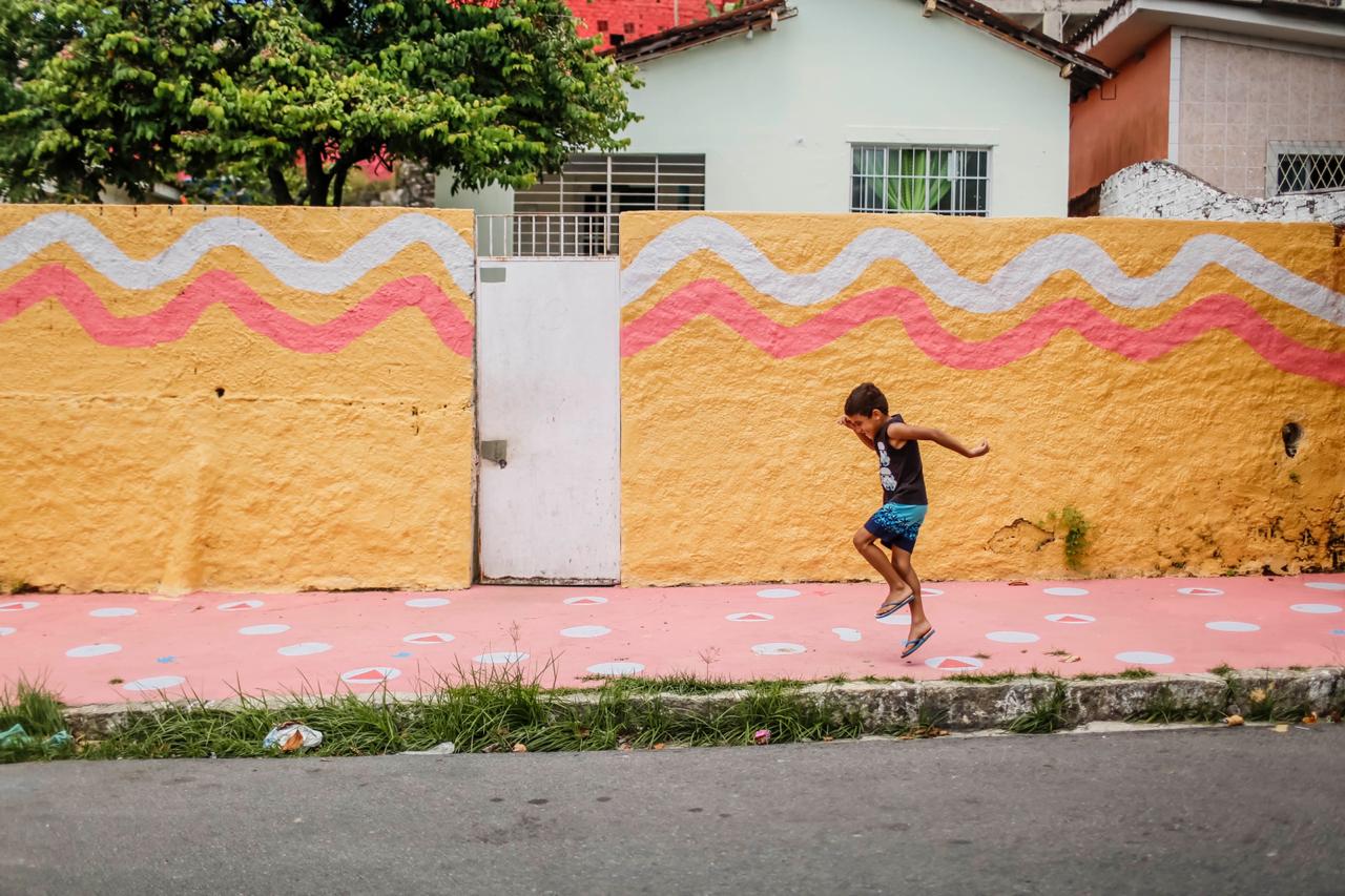 Muro colorido de uma casa, no Vasco da Gama. Menino pula na frente do muro, em uma calçada com pegadas desenhadas.