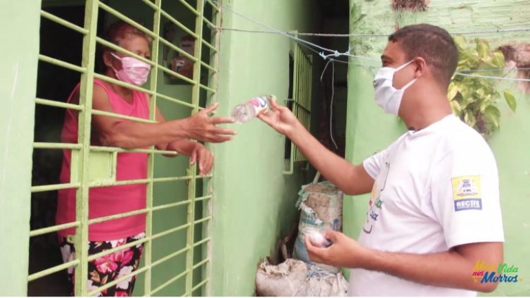 Mulher está dentro de casa, usando máscara. Pelo portão, ela recebe itens de higiene de um voluntário.